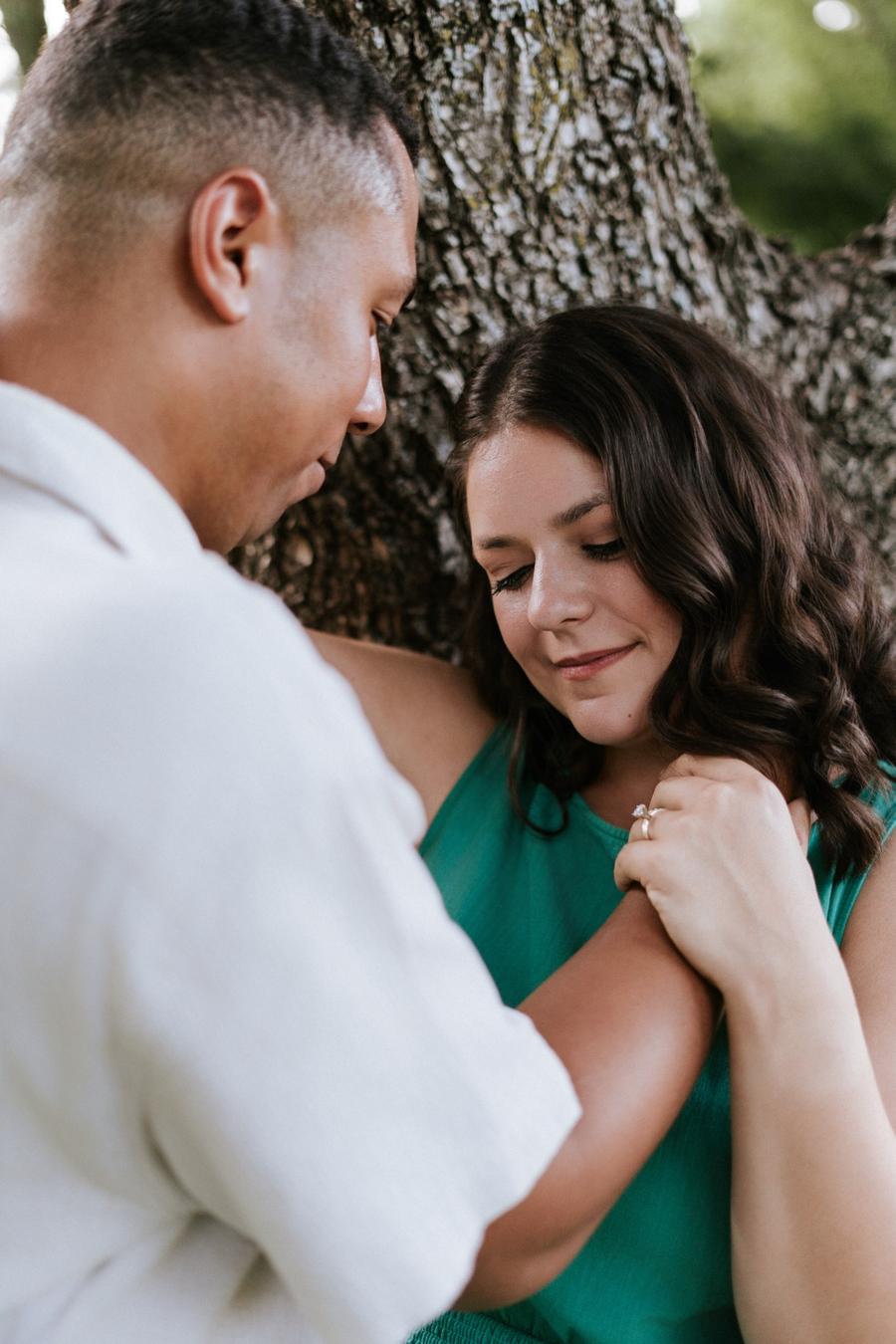 Engaged couple sharing a quiet, intimate moment beneath a tree during their engagement session, with the bride-to-be showing off her ring.