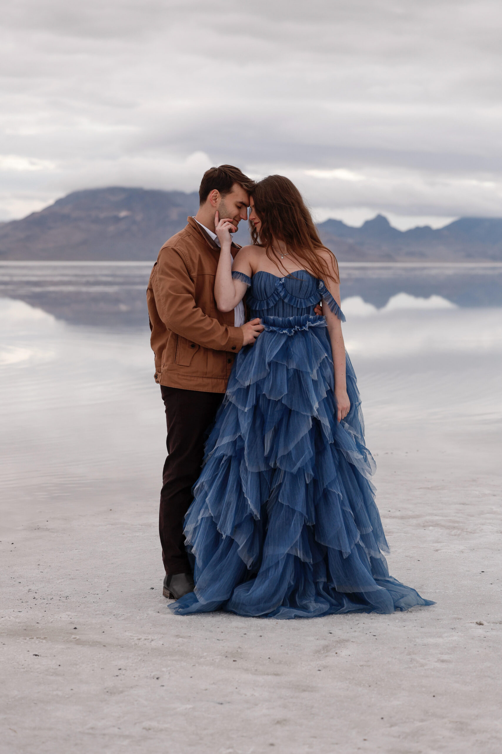 A couple shares an intimate moment standing on a reflective salt flat, with dramatic mountains and a cloudy sky in the background. The bride wears a layered dusty blue gown while the groom wears a warm brown jacket, creating a striking contrast against the soft neutral landscape.