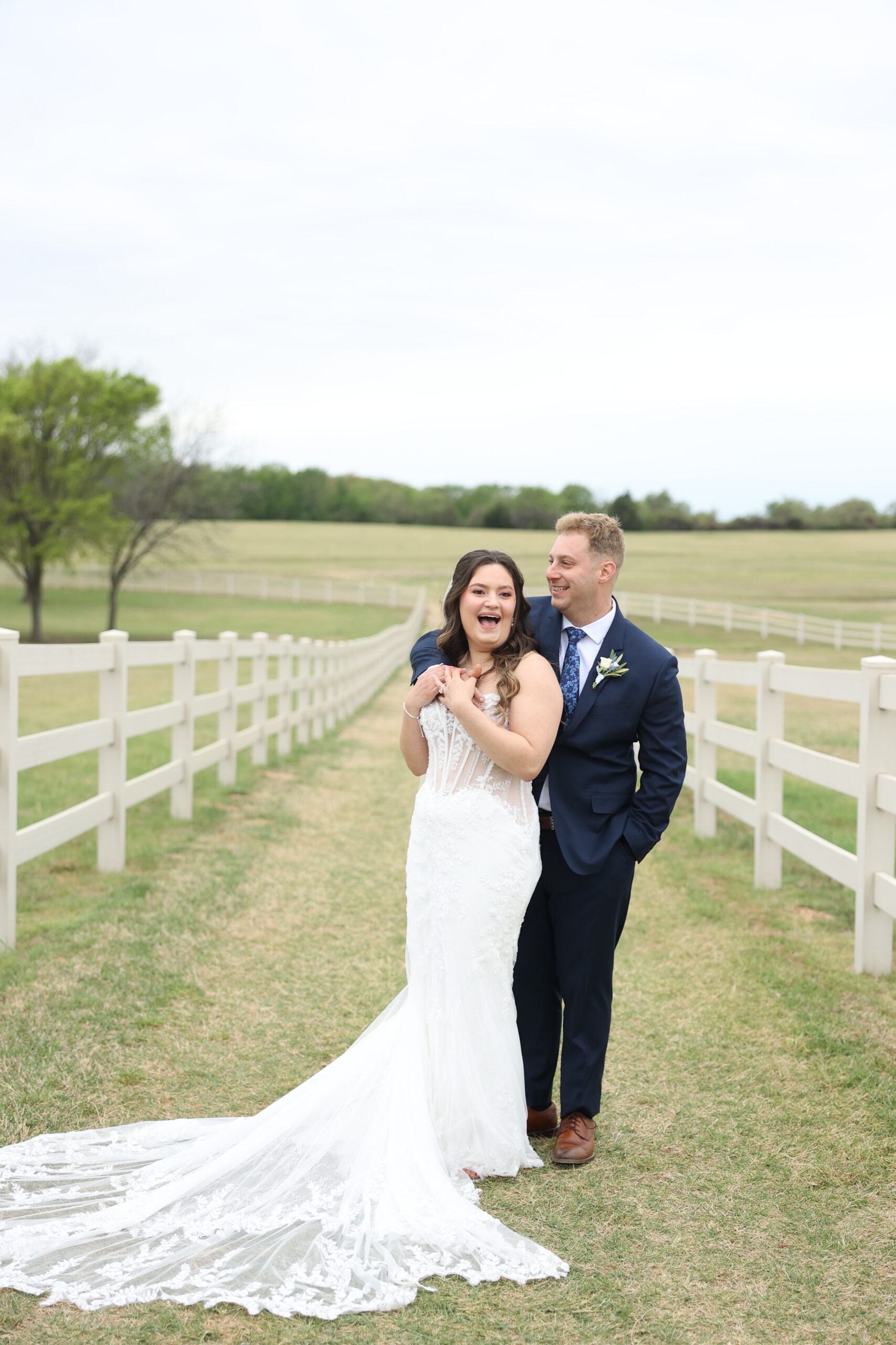 Bride and groom standing together in a grassy pathway at Bella Cavalli Events, surrounded by white fencing and open countryside, sharing a joyful moment on their wedding day.